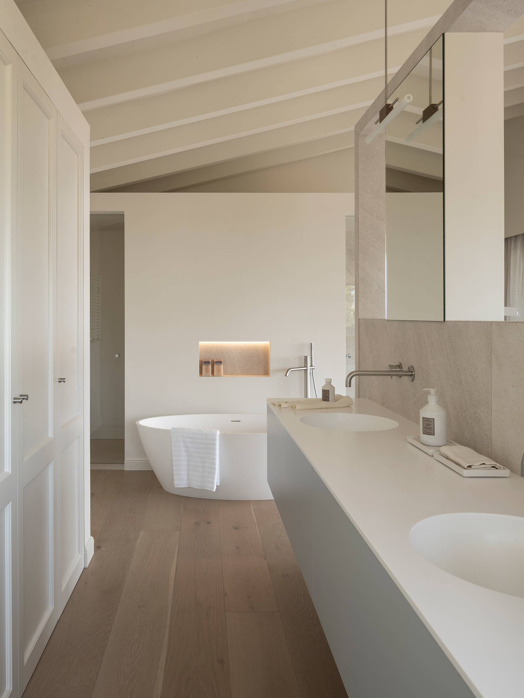 Baño con bañera exenta, doble lavabo sobre encimera blanca, revestimiento efecto piedra y suelo de madera