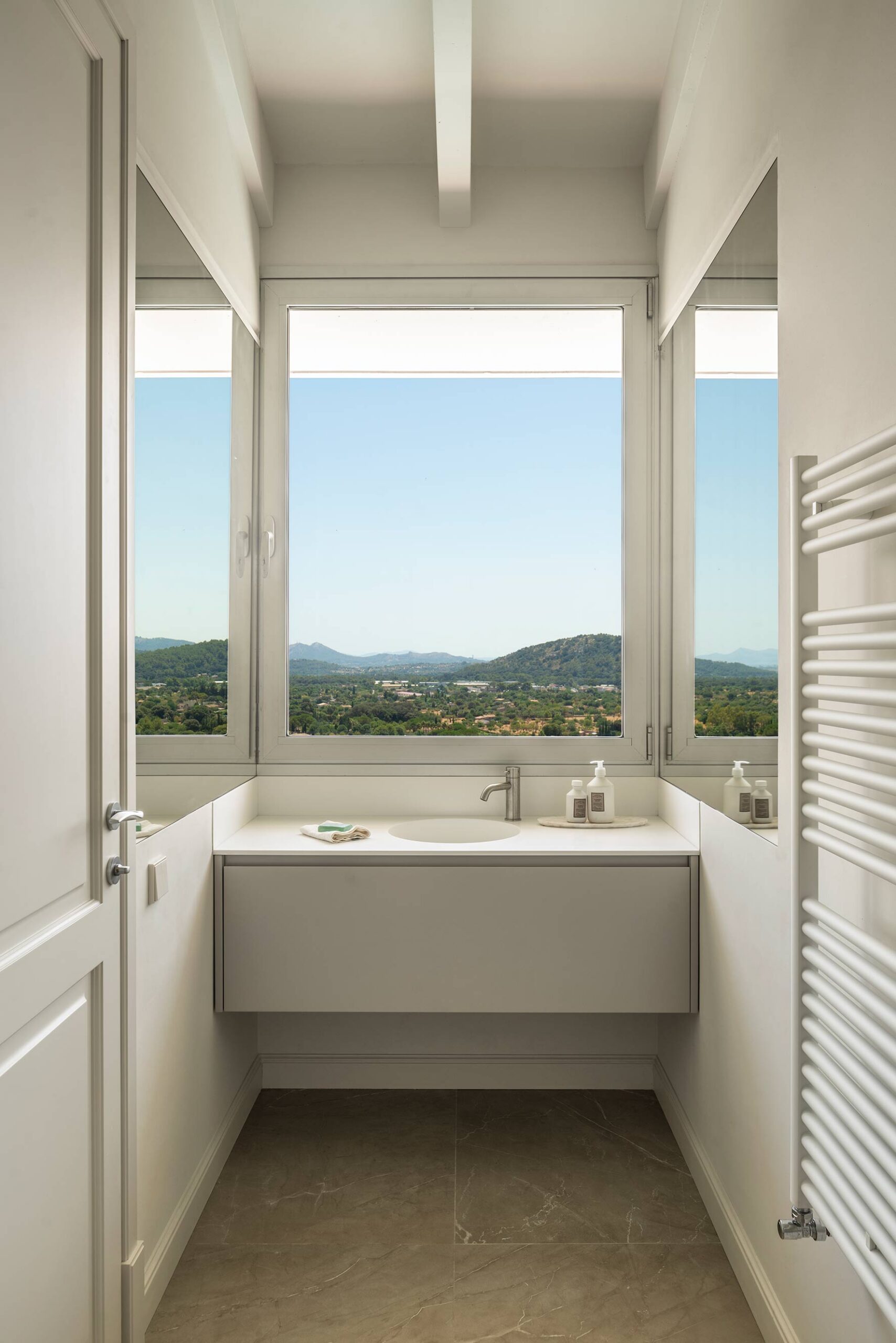 Baño con lavabo suspendido bajo una ventana con vistas a las montañas y radiador toallero blanco