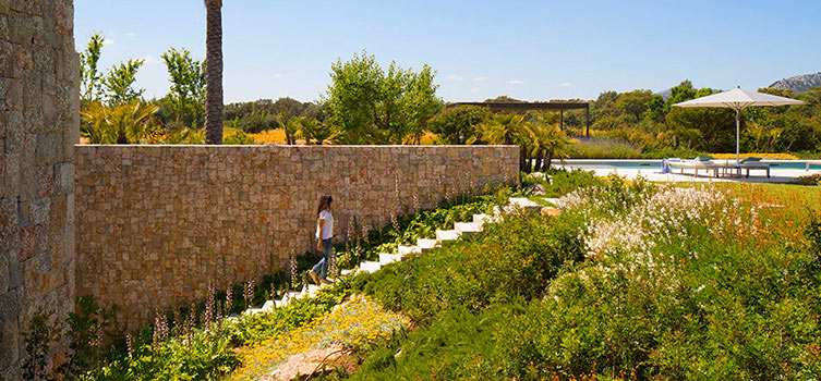 Escalera de piedra clara entre arbustos y flores junto a un muro de piedra, con piscina al fondo