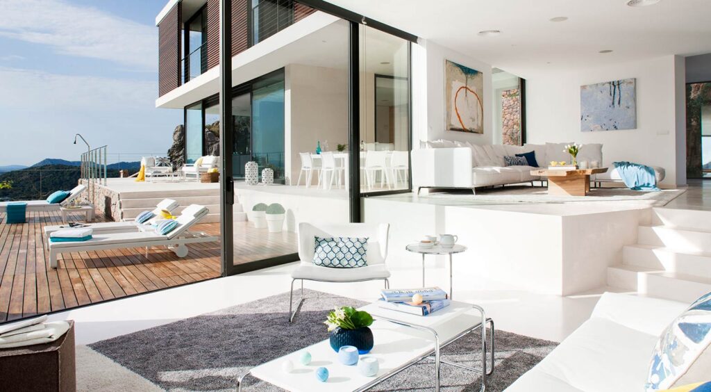 Zur Terrasse geöffnetes Wohnzimmer mit weißen Sofas, Couchtisch und Sonnenliegen auf Holzdeck