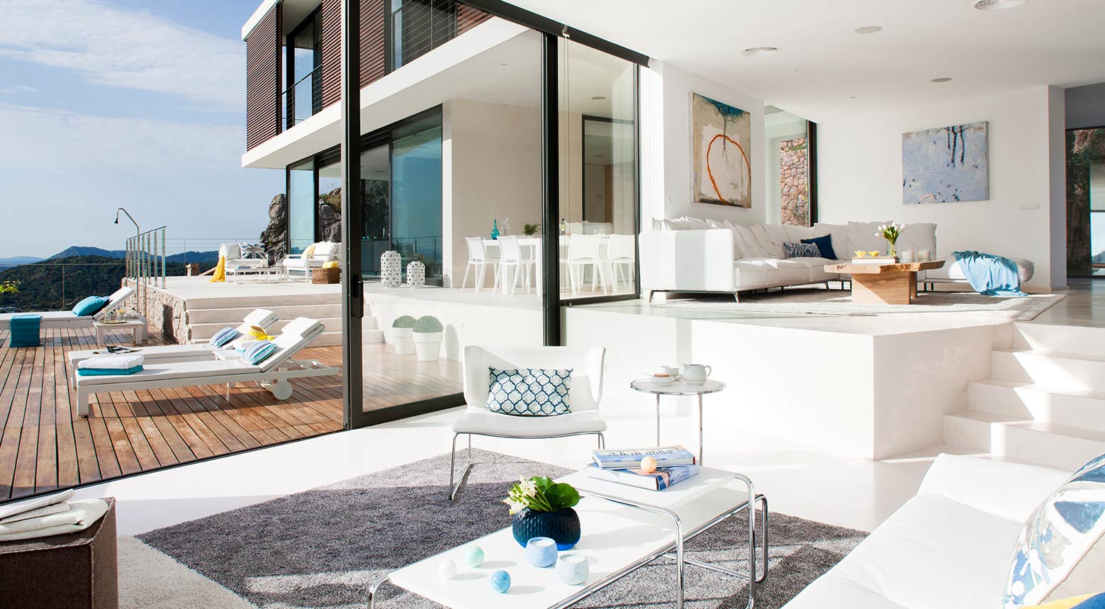 Zur Terrasse geöffnetes Wohnzimmer mit weißen Sofas, Couchtisch und Sonnenliegen auf Holzdeck