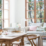 Comedor con mesa y sillas de madera de asiento trenzado frente al salón y ventanales con palmeras