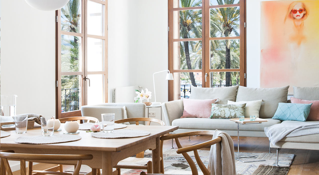 Comedor con mesa y sillas de madera de asiento trenzado frente al salón y ventanales con palmeras