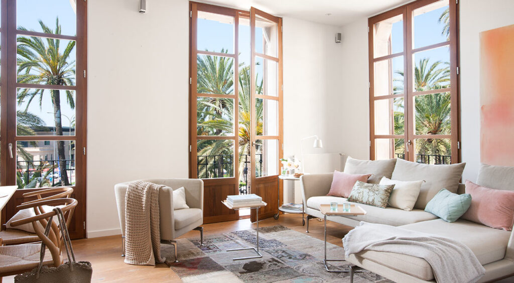 Salón del piso Antoni Maura con ventanales de madera, vistas a palmeras y sofá claro