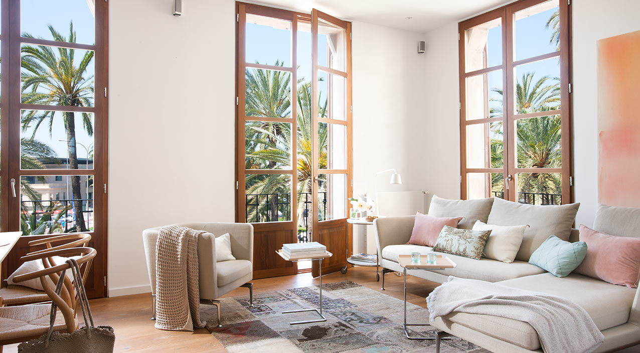 Salón del piso Antoni Maura con ventanales de madera, vistas a palmeras y sofá claro