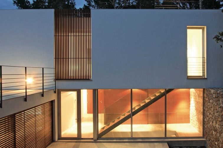 Weiße Fassade von Blanca pureza in der Abenddämmerung mit Holzlamellen, beleuchteter Verglasung und Steinmauer