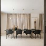 Comedor con mesa larga, sillas negras, lámpara lineal dorada y cortinas de lino entre lamas verticales