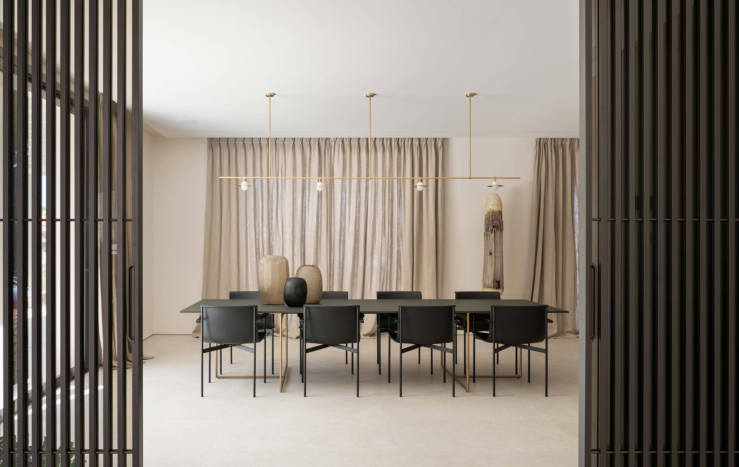 Comedor con mesa larga, sillas negras, lámpara lineal dorada y cortinas de lino entre lamas verticales