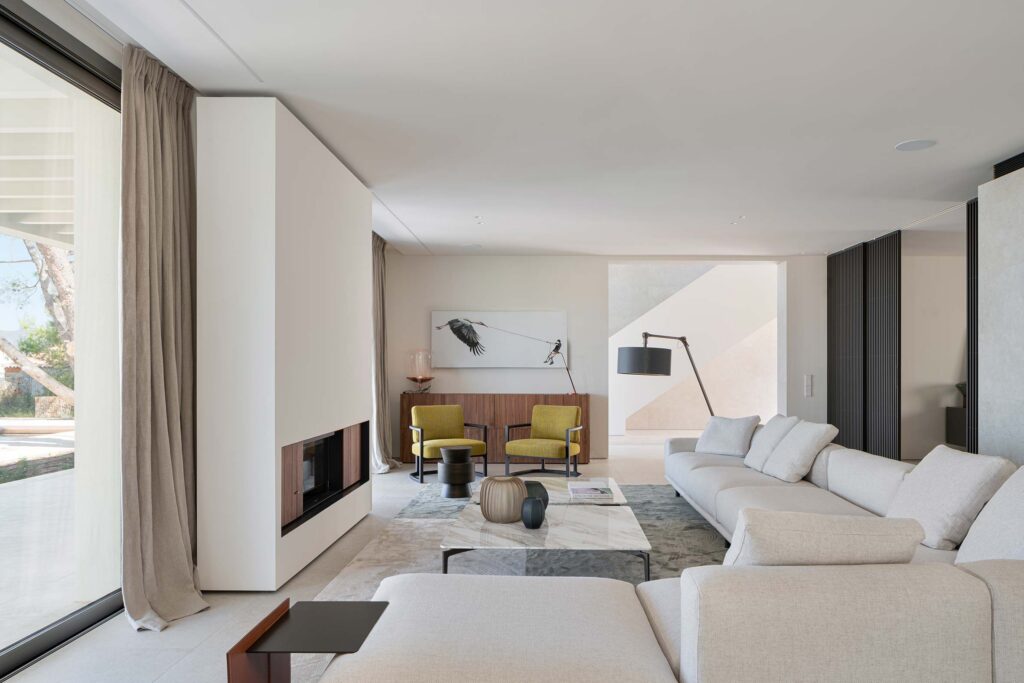 Salón con sofá crema, chimenea revestida, mesa de mármol, butacas mostaza y gran ventanal