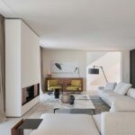 Salón con sofá crema, chimenea revestida, mesa de mármol, butacas mostaza y gran ventanal