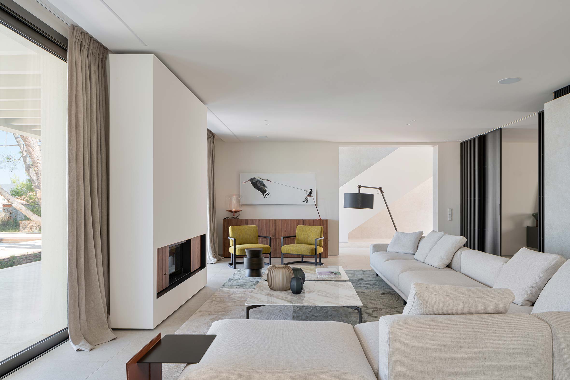 Salón con sofá crema, chimenea revestida, mesa de mármol, butacas mostaza y gran ventanal
