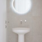 Lavabo de pie blanco con espejo redondo retroiluminado y grifería empotrada sobre revestimiento claro