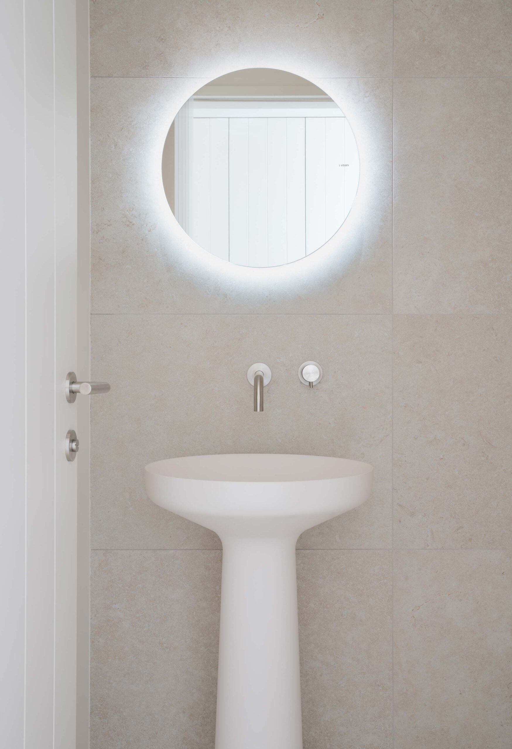 Lavabo de pie blanco con espejo redondo retroiluminado y grifería empotrada sobre revestimiento claro