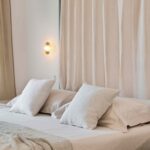 Detalle de cama baja con almohadones blancos, manta tejida, aplique dorado y cortina translúcida