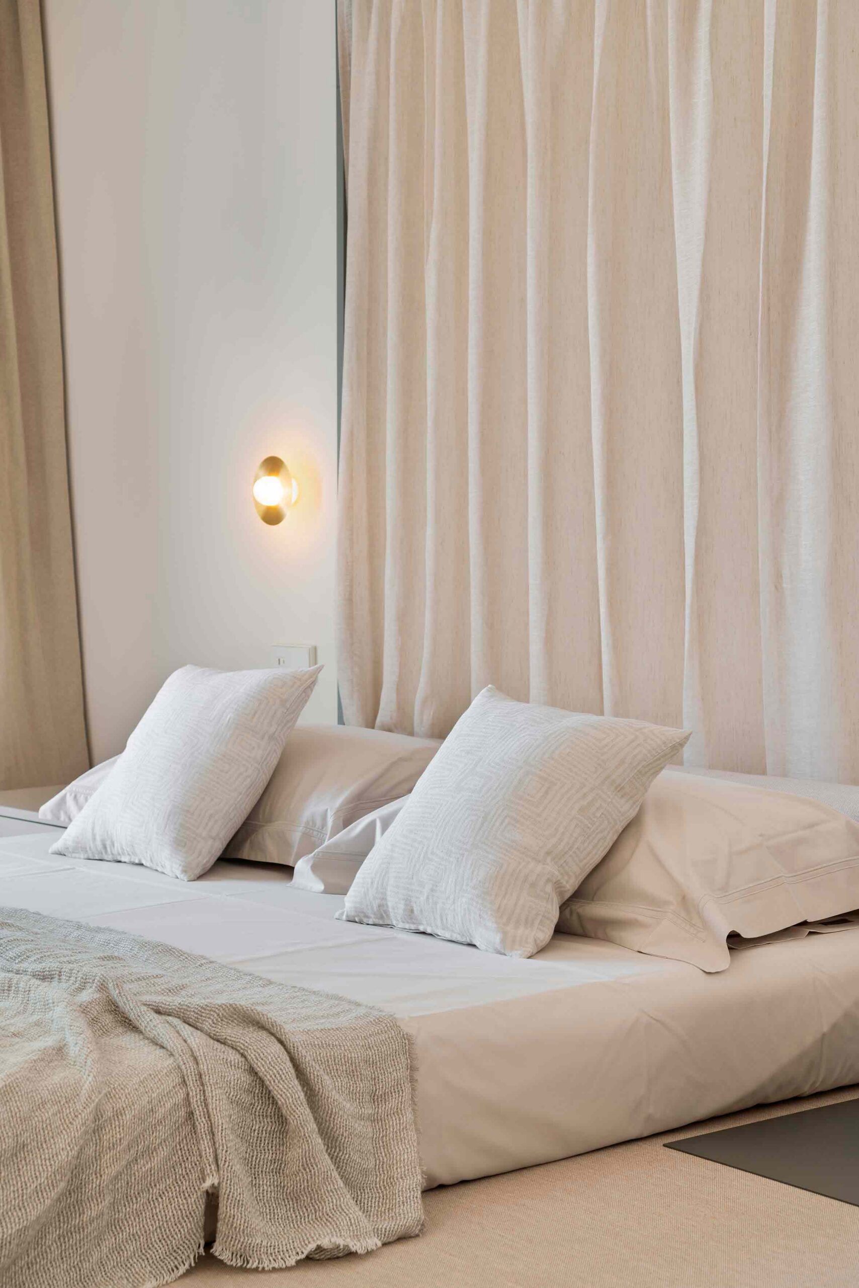Detalle de cama baja con almohadones blancos, manta tejida, aplique dorado y cortina translúcida