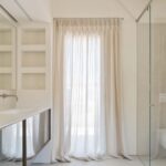 Baño con lavabo doble blanco, espejo, nichos empotrados, cortina translúcida y mampara de cristal