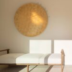 Diván de madera con cojines blancos bajo un disco dorado decorativo, sobre suelo moteado claro