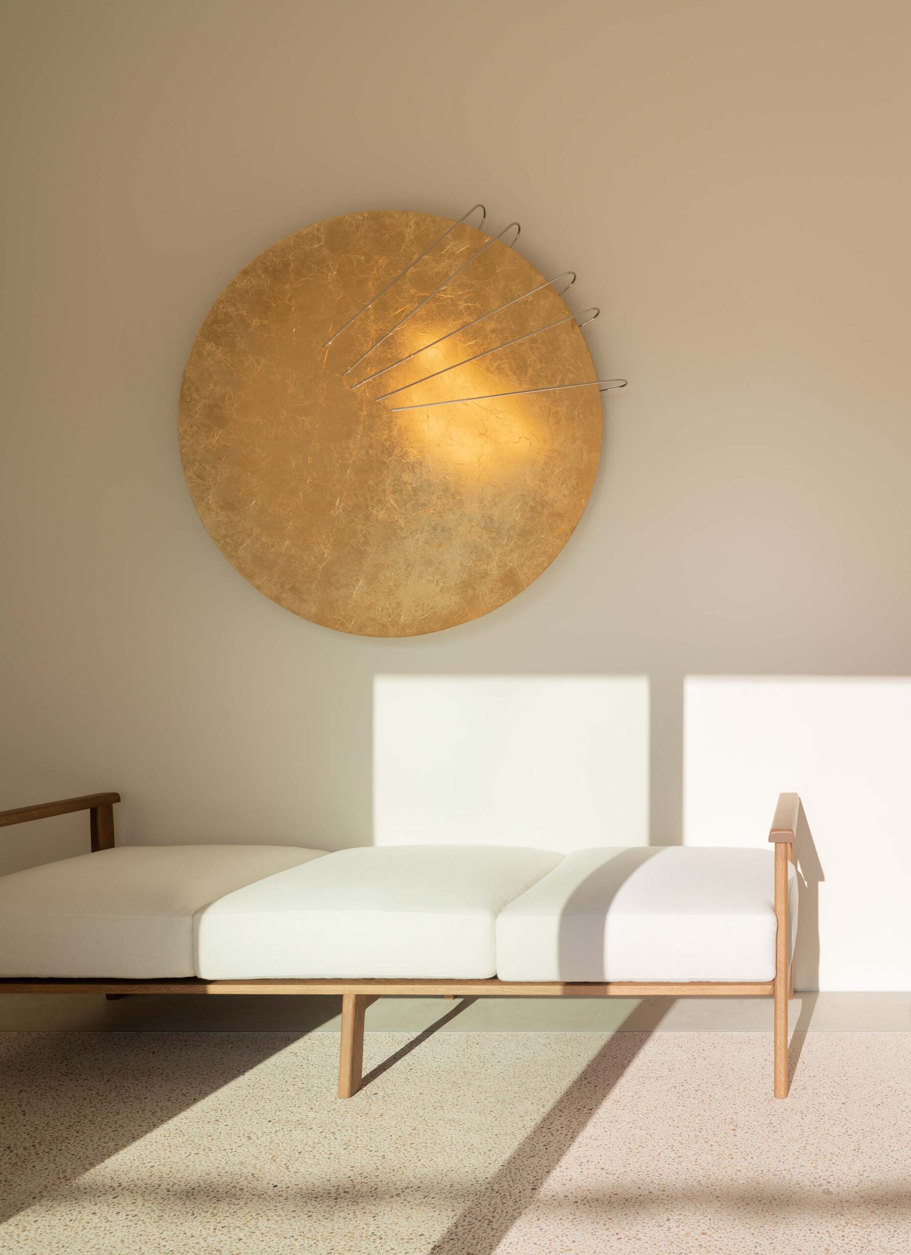 Diván de madera con cojines blancos bajo un disco dorado decorativo, sobre suelo moteado claro
