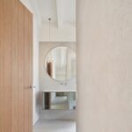 Pasillo hacia el baño con puerta de madera, espejo redondo, mueble suspendido y lámpara colgante