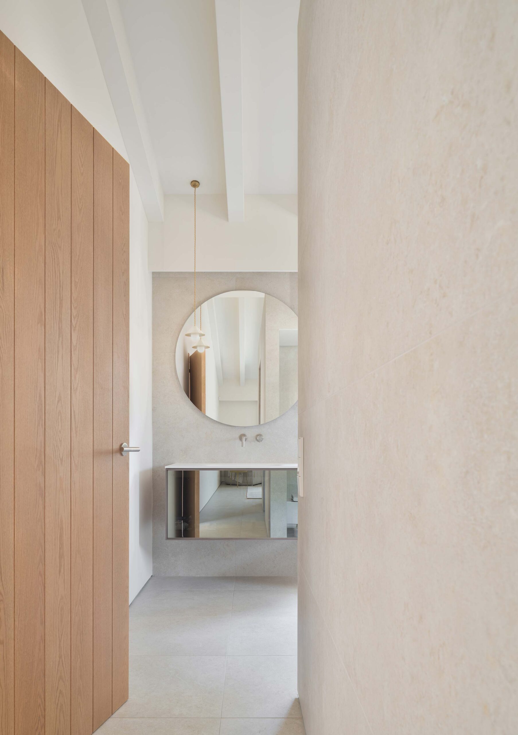 Pasillo hacia el baño con puerta de madera, espejo redondo, mueble suspendido y lámpara colgante