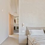 Dormitorio con muro claro, mesita gris y puerta de madera abierta al baño con espejo redondo