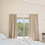 Dormitorio con cortinas de lino, puerta al balcón con vistas a pinos y tocador negro con espejo