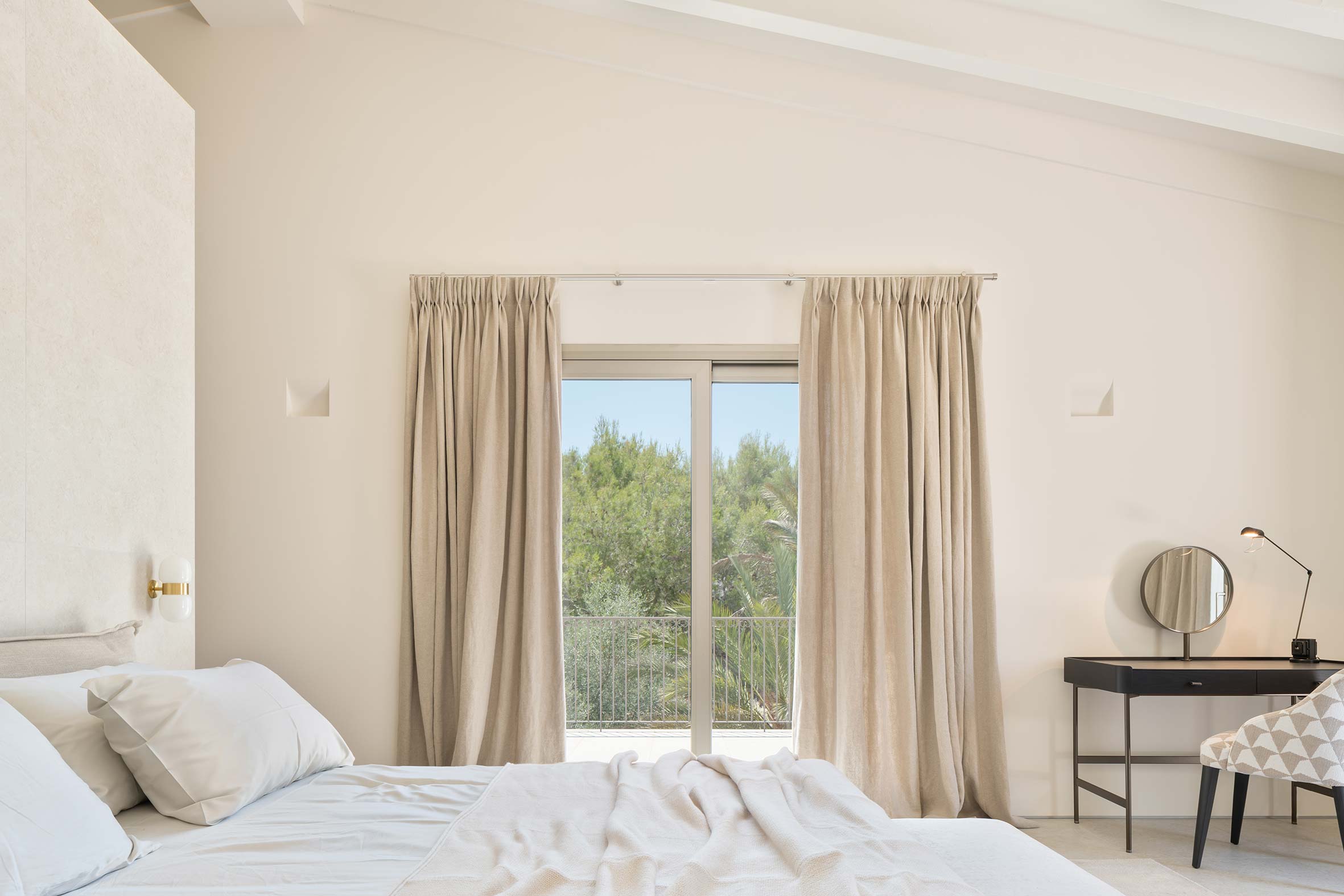 Dormitorio con cortinas de lino, puerta al balcón con vistas a pinos y tocador negro con espejo