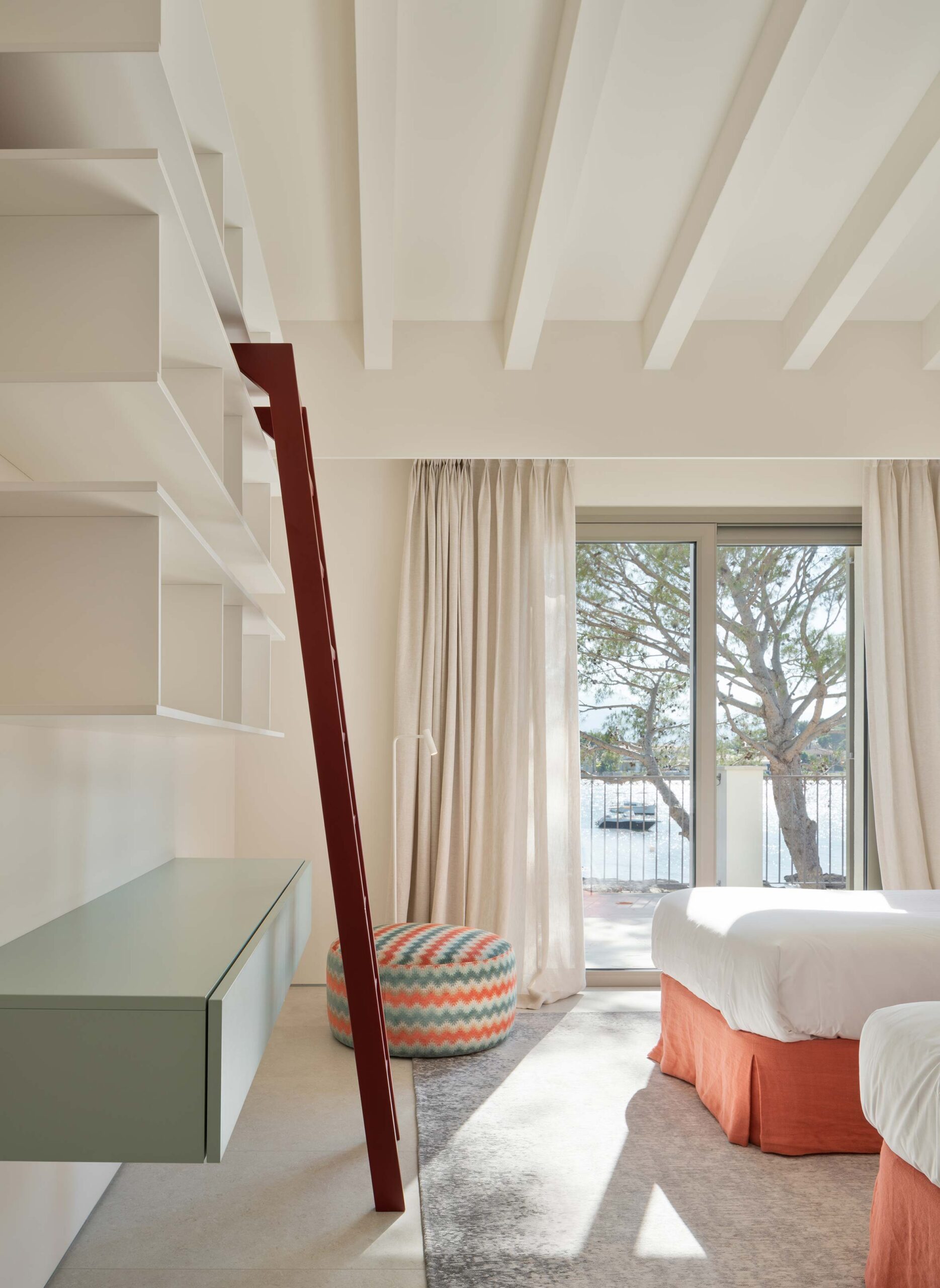 Dormitorio con dos camas de faldón naranja, estantería con escalera roja y vistas a la bahía