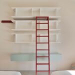 Estantería blanca con escalera roja y mueble suspendido verde agua entre dos camas