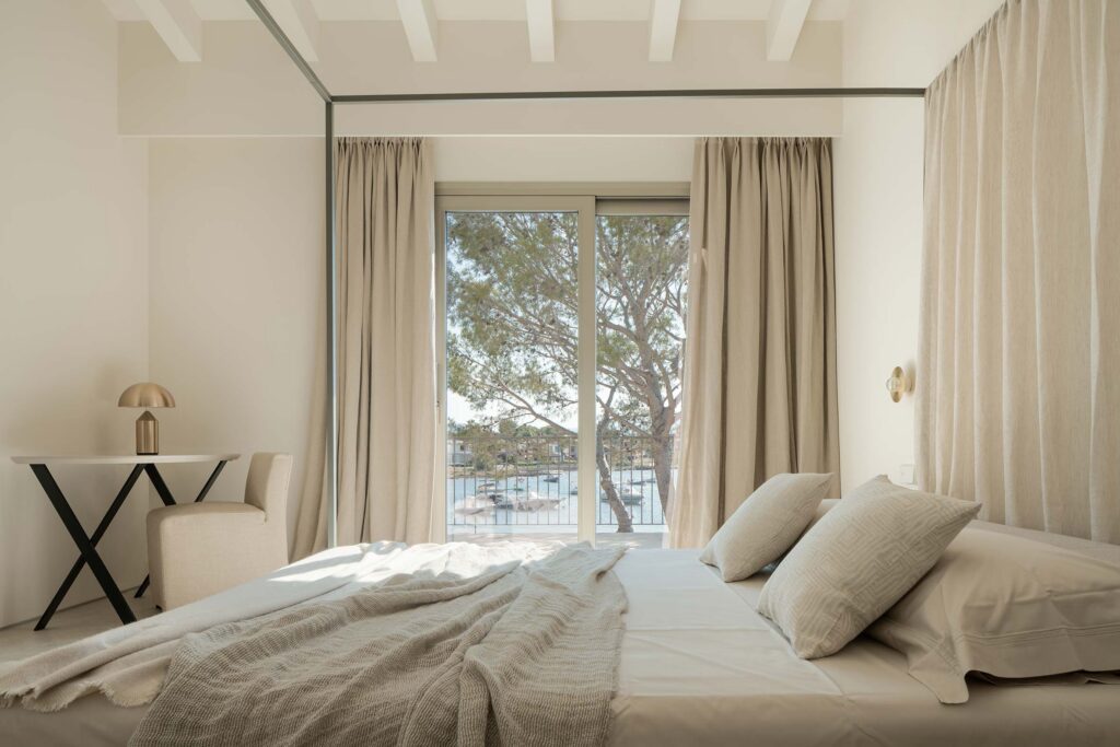 Dormitorio con cama de dosel, cortinas de lino beige y vistas a una bahía con barcas