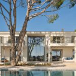 Fachada con cristalera de doble altura, balcones con contraventanas, un pino y la piscina reflejando la casa