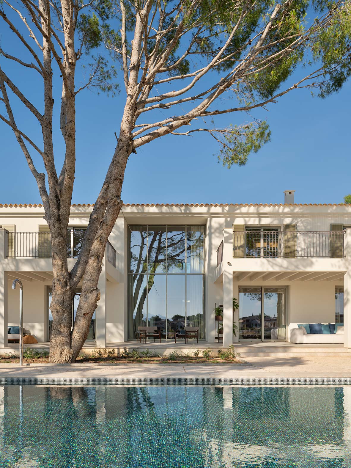 Fachada con cristalera de doble altura, balcones con contraventanas, un pino y la piscina reflejando la casa