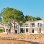 Villa Voramar, fachada blanca de dos plantas con muro de piedra y pinos junto al agua