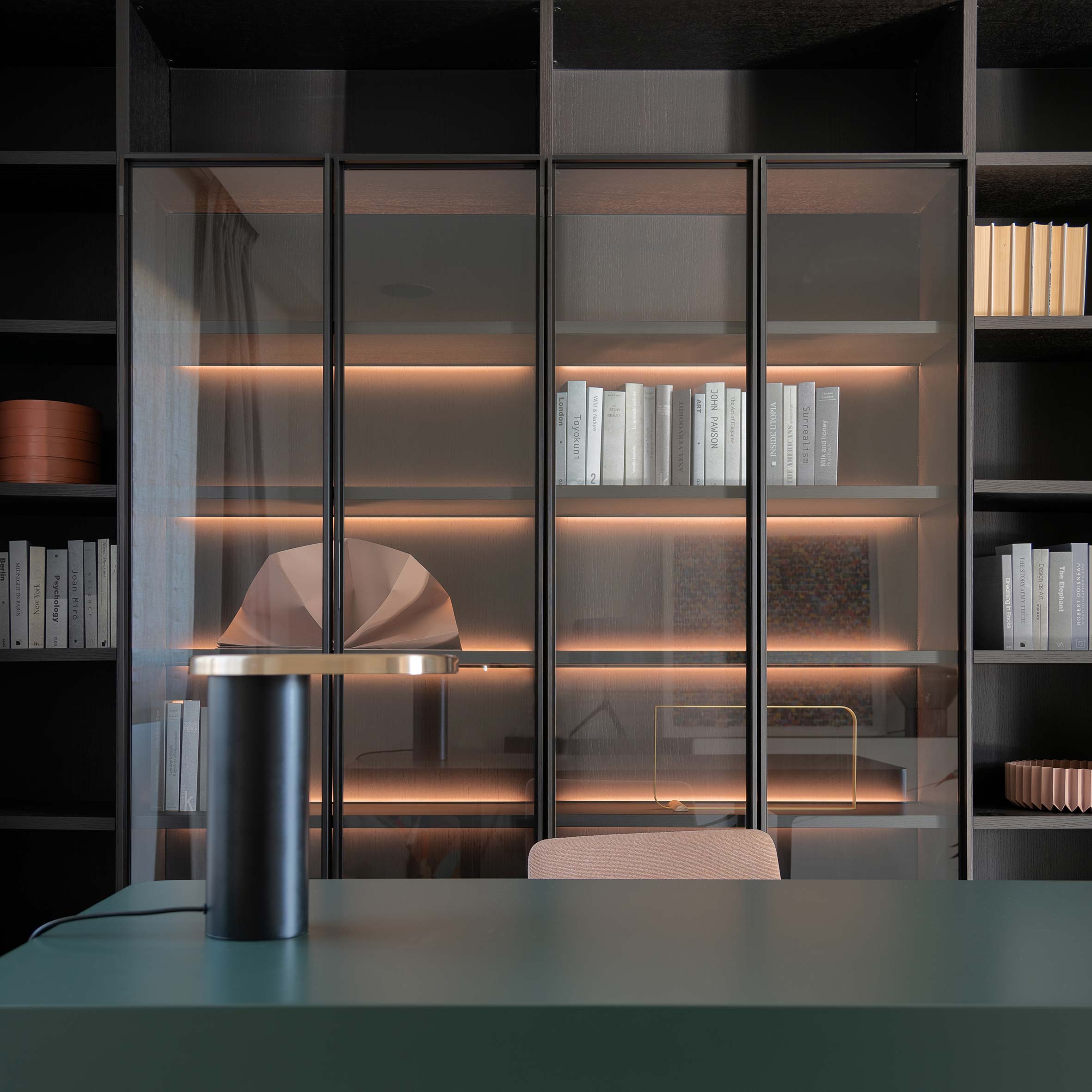 Biblioteca de madera oscura con vitrinas de cristal iluminadas, escritorio verde y lámpara negra