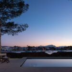 Piscina al anochecer frente a la bahía con montañas al fondo, pino y sillas de tijera