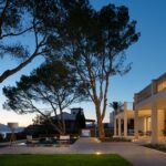 Jardín y porche iluminados al atardecer, con pinos, césped, sillas de tijera y luz cálida