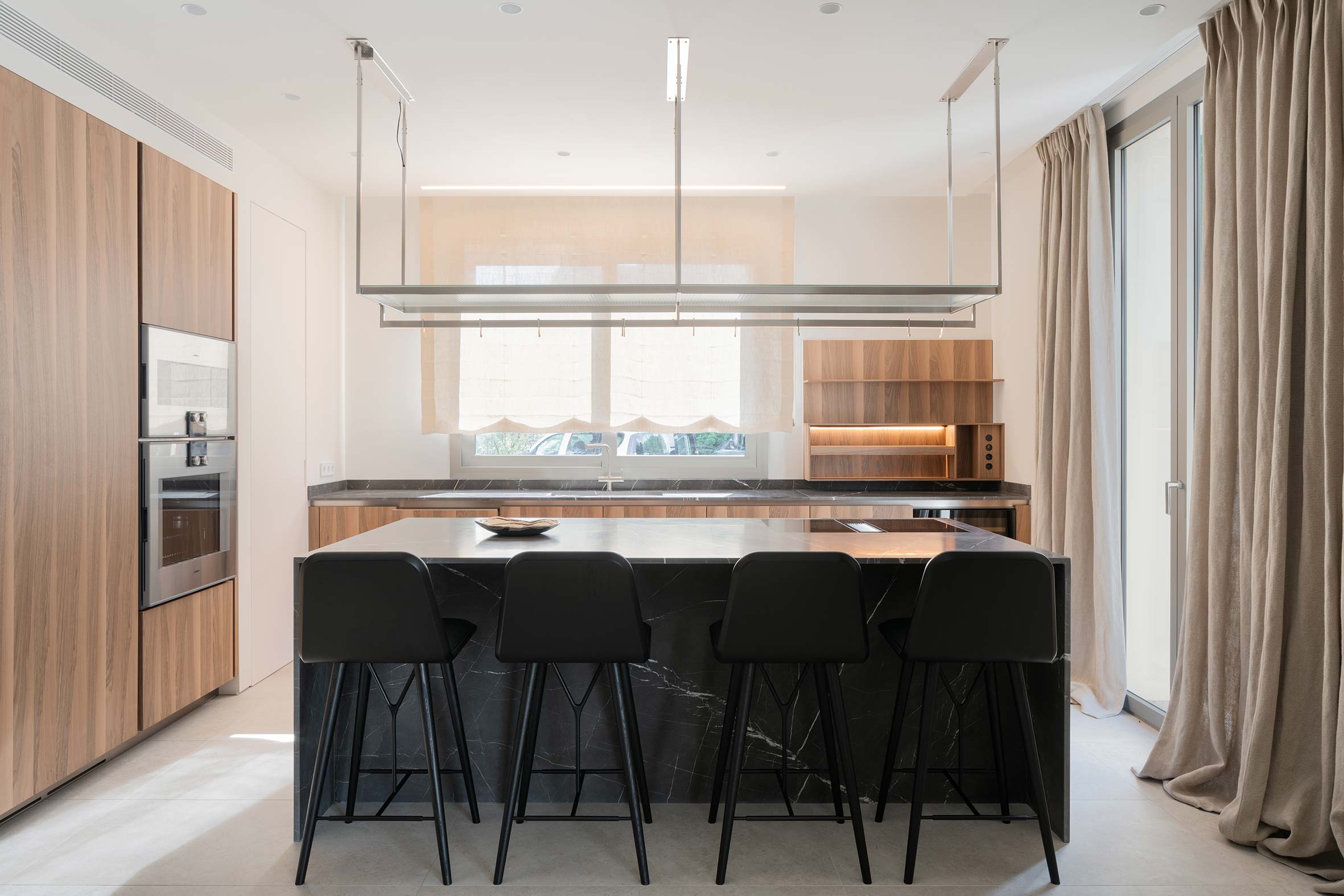 Cocina con isla de mármol oscuro, cuatro taburetes negros, armarios de madera y estante colgante