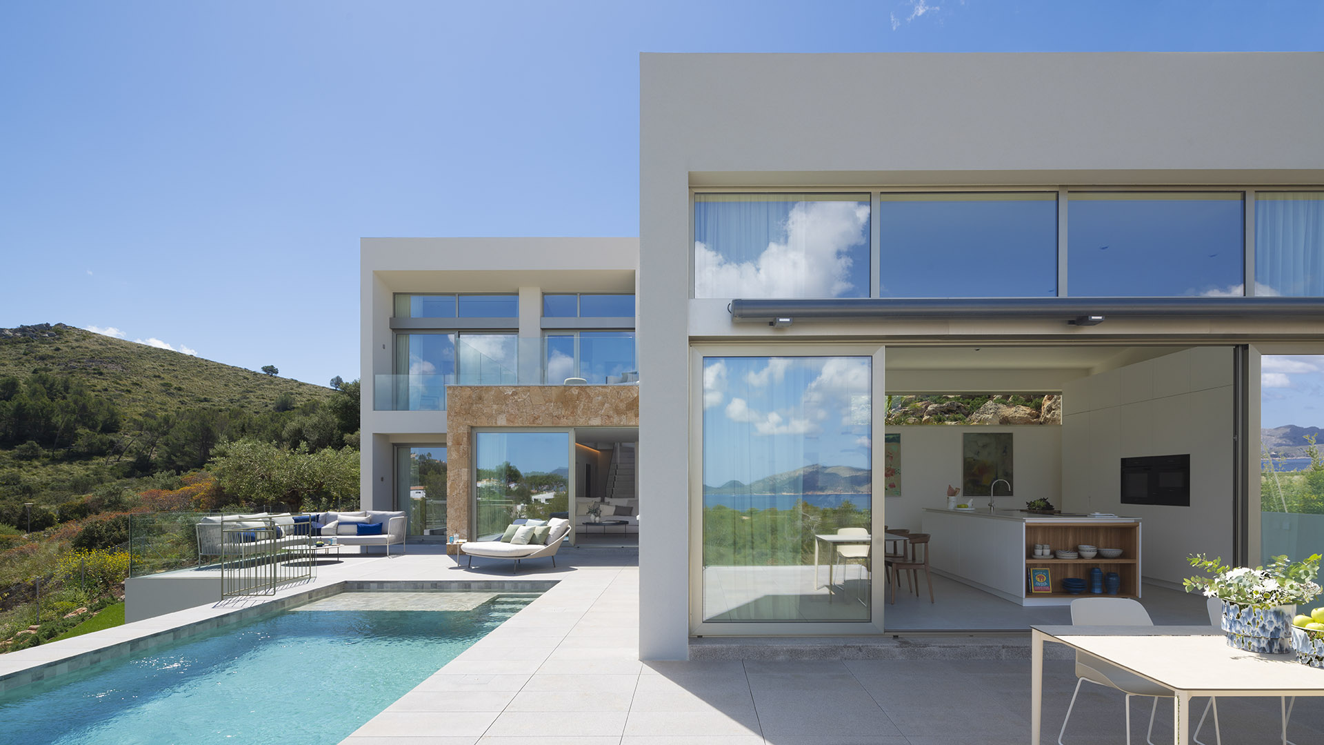 Terraza de pavimento claro junto a la piscina, con cocina abierta al exterior y ladera verde