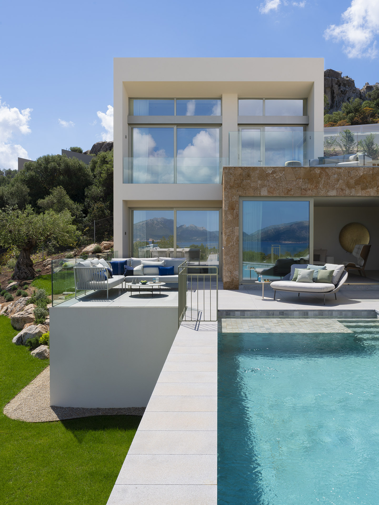 Terrazas escalonadas de la villa con sofás exteriores, piedra natural, césped, olivo y piscina turquesa