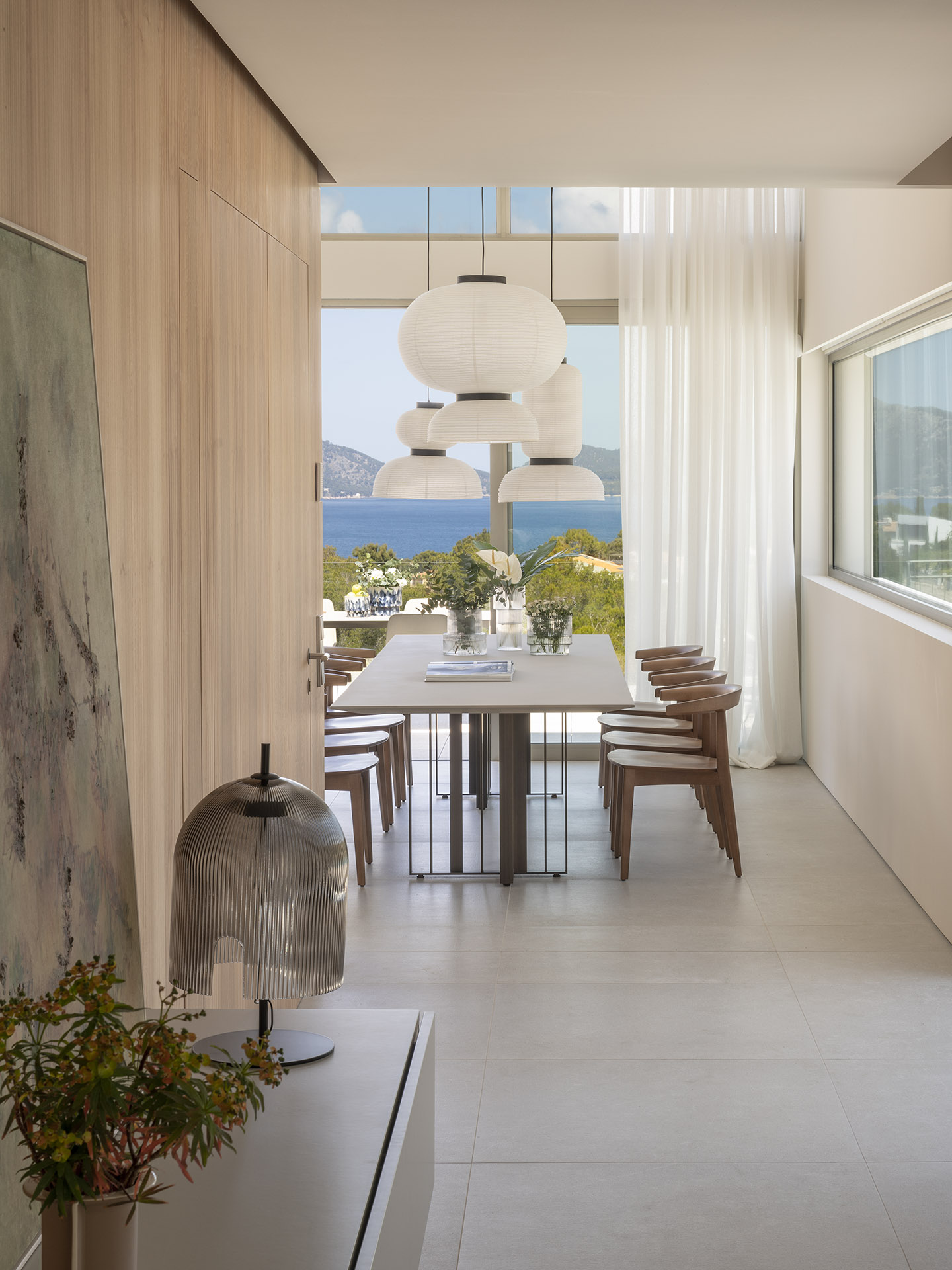 Comedor de doble altura con mesa alargada, sillas de madera, lámparas de papel y mar al fondo