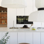 Detalle de la cocina con lámparas de papel, armarios blancos, hornos negros y estantería de madera