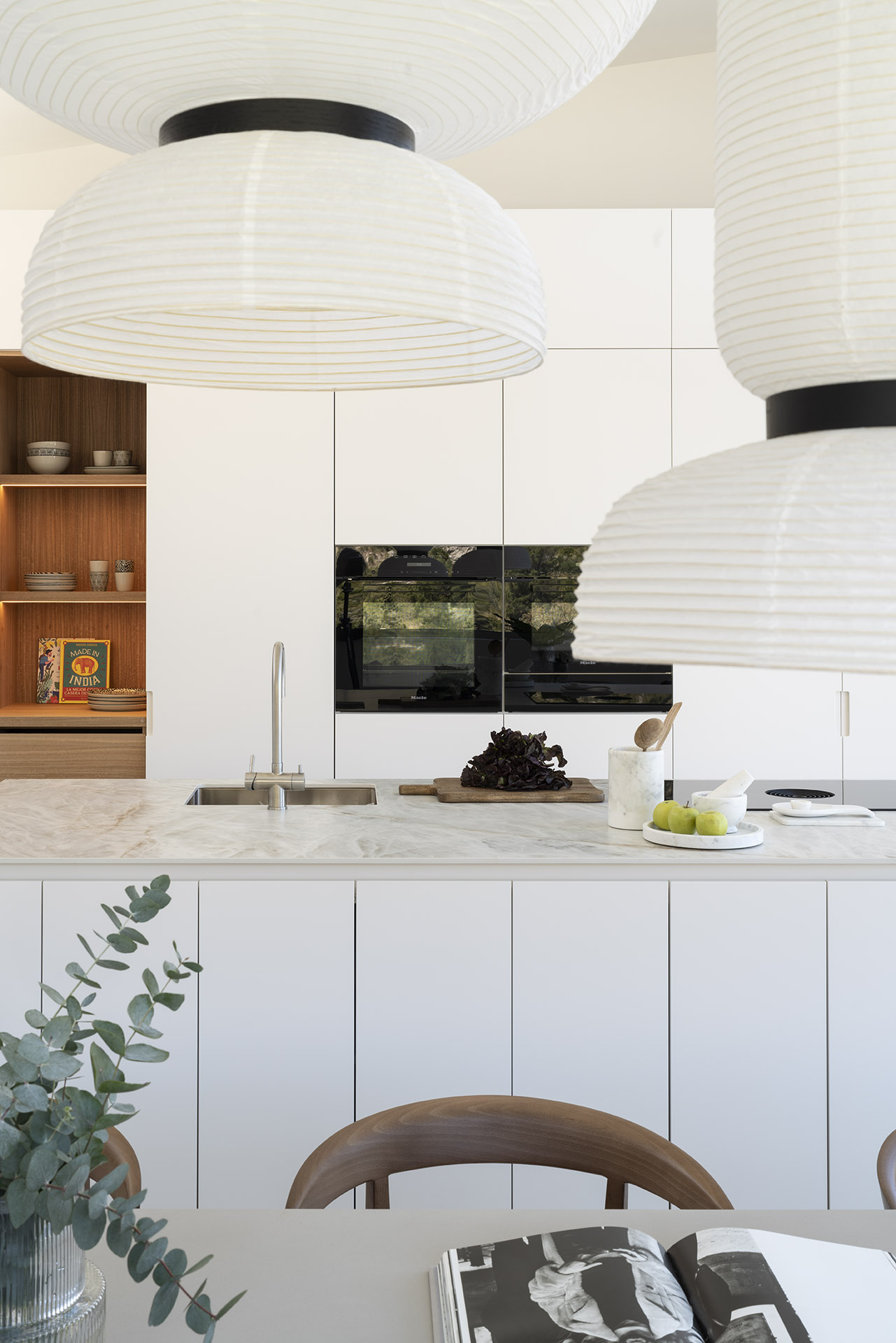 Detalle de la cocina con lámparas de papel, armarios blancos, hornos negros y estantería de madera