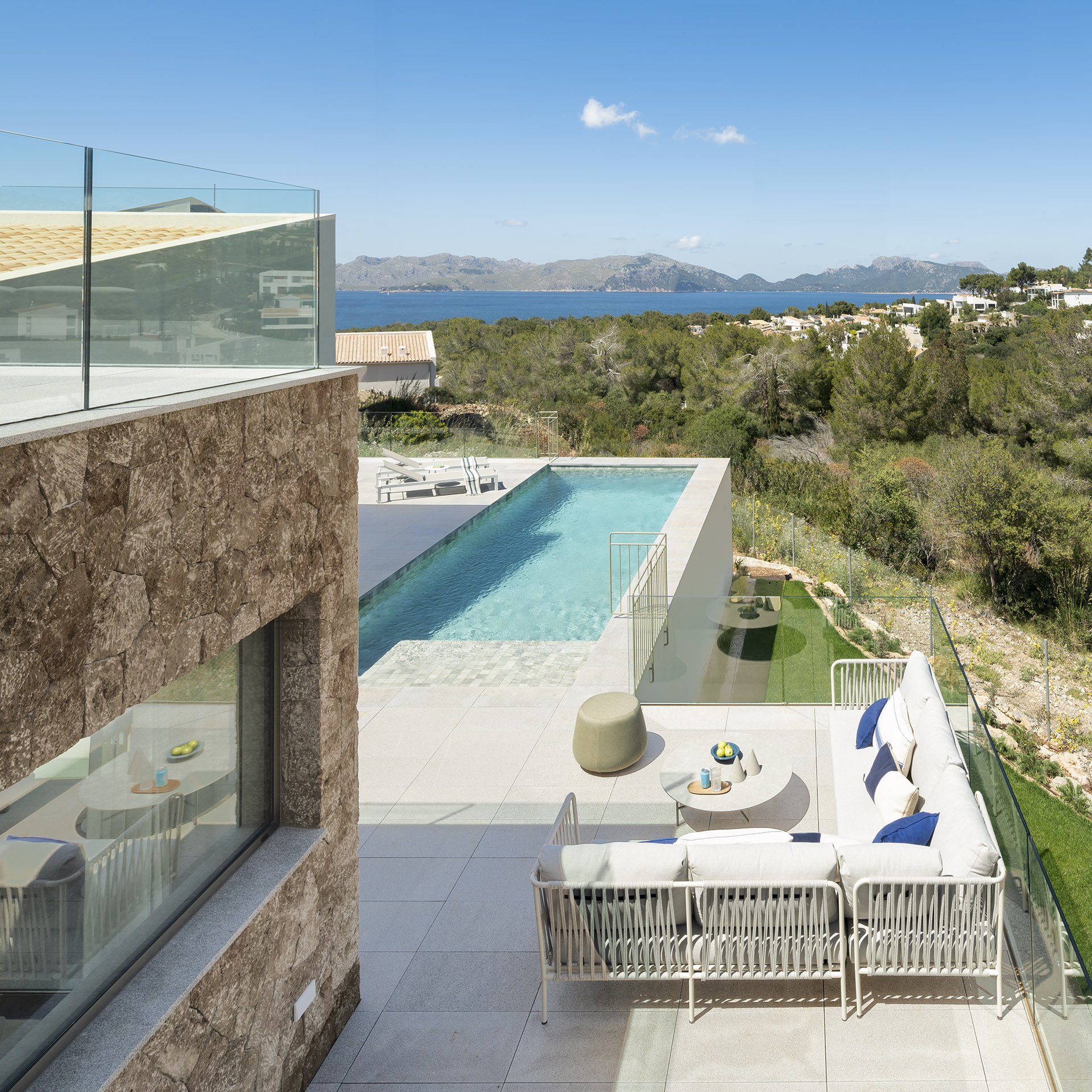 Vista elevada de la piscina alargada, zona de estar exterior, muro de piedra, mar y montañas