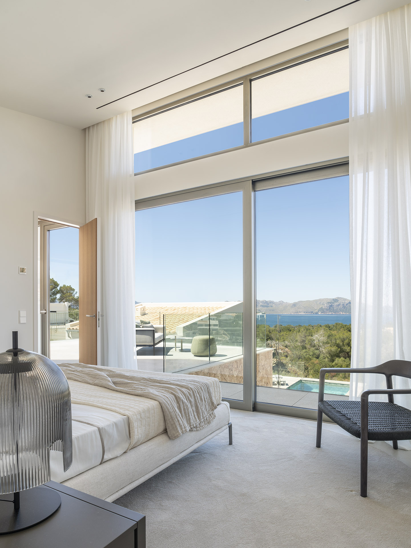 Dormitorio con ventanal de doble altura, cortinas blancas y vistas al mar desde la cama