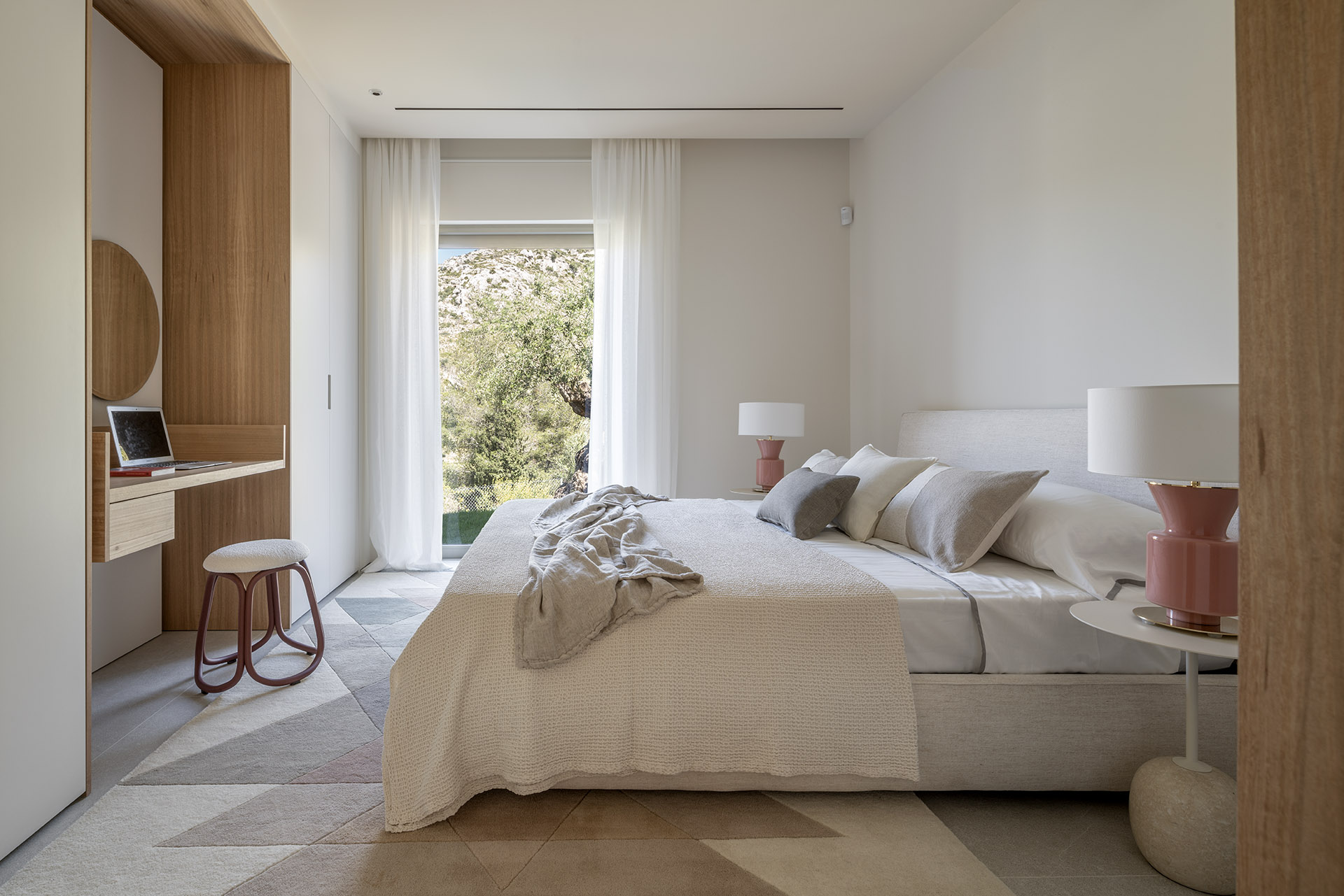 Dormitorio con cama doble, escritorio integrado en madera, alfombra geométrica y ventanal al jardín