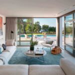 Salón con sofá claro, alfombra turquesa, cuadro abstracto y cristaleras abiertas a la piscina
