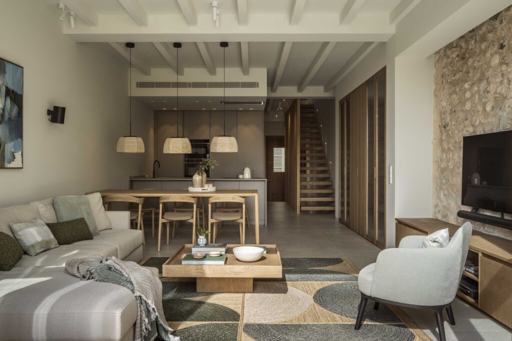 Open-plan living area with sofa, dining area, kitchen at the back, wooden staircase and stone wall