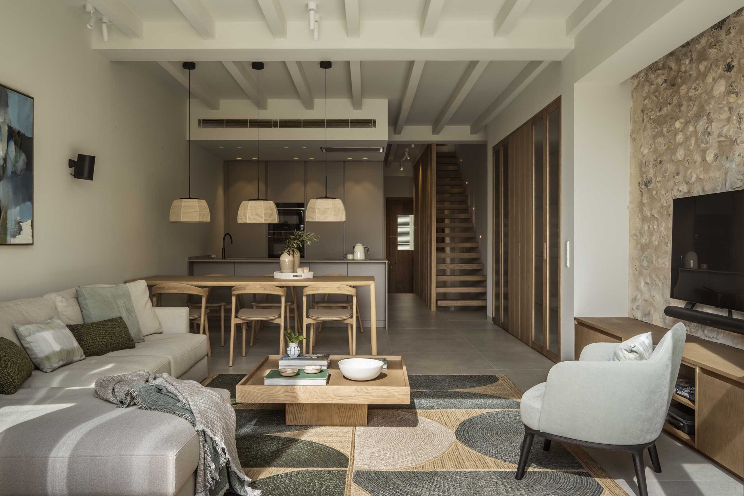 Open-plan living area with sofa, dining area, kitchen at the back, wooden staircase and stone wall
