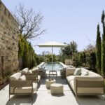 Outdoor lounge with light upholstered armchairs and sofas, white hexagonal side tables and cypress trees