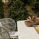 Detail of an outdoor table with light tabletop, wooden bowls, rope chairs and fuchsia flowers